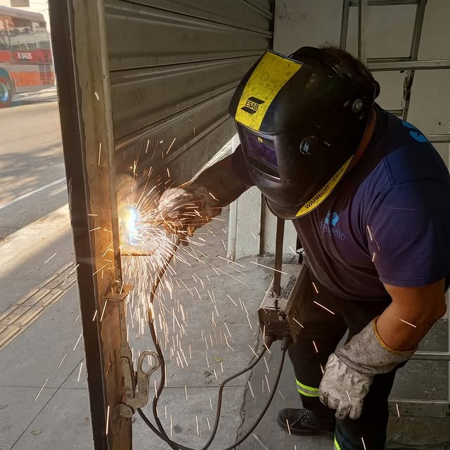 Técnico da Maranata soldando a estrutura de uma porta de aço na calçada, com faíscas do maçarico
